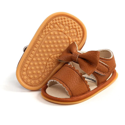 A cute pair of Bubebaby BabyLove  faux leather sandals for infants featuring an adorable bow detail. The sandals are designed for comfort and style, perfect for little ones to wear during warm weather. The adjustable strap ensures a secure fit while the soft sole provides cushioning for tiny feet